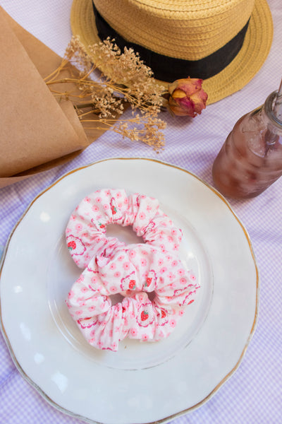 two pink scrunchies with a flower, strawberry, and heart print on a plate in a picnic setting