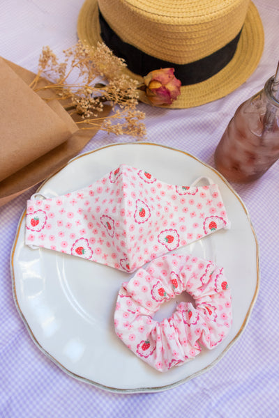 a pink scrunchie with a flower, strawberry, and heart print. matching mask sits above the scrunchie on a plate in a picnic setting