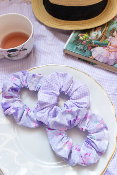 a purple gingham scrunchie and two purple scrunchies placed on plate in a picnic setting