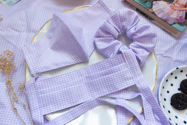 a purple and white gingham print scrunchie. matching pleated mask and non-pleated mask sit beside the scrunchie on a plate in a picnic setting.