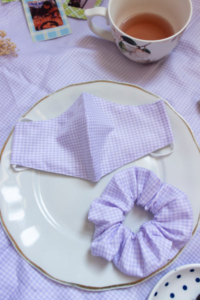 a purple and white gingham print scrunchie. matching mask sits above the scrunchie on a plate in a picnic setting.