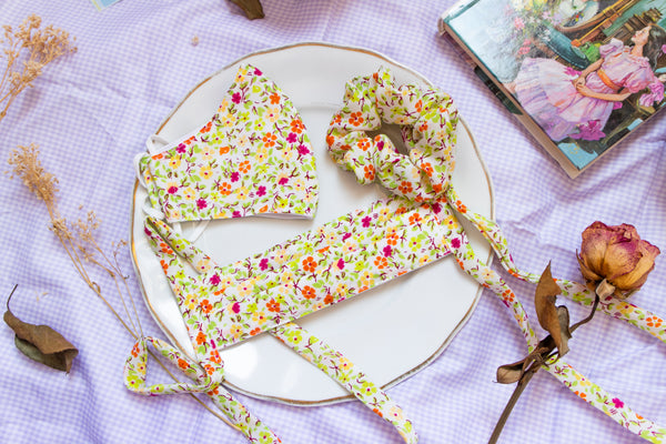 a warm toned scrunchie reminiscent of spring with a green, yellow, orange, and magenta flower print. matching pleated mask and non-pleated mask sit beside the scrunchie on a plate in a picnic setting.