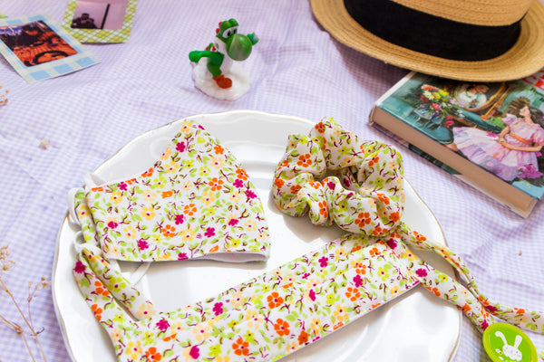 a warm toned scrunchie reminiscent of spring with a green, yellow, orange, and magenta flower print. matching pleated mask and non-pleated mask sit beside the scrunchie on a plate in a picnic setting.