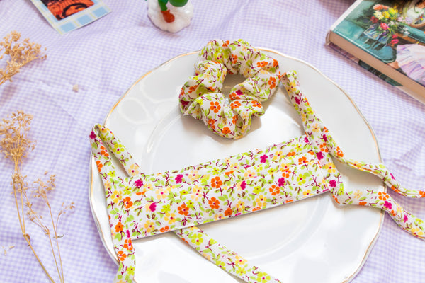 a warm toned scrunchie reminiscent of spring with a green, yellow, orange, and magenta flower print. matching pleated mask sits below the scrunchie on a plate in a picnic setting.